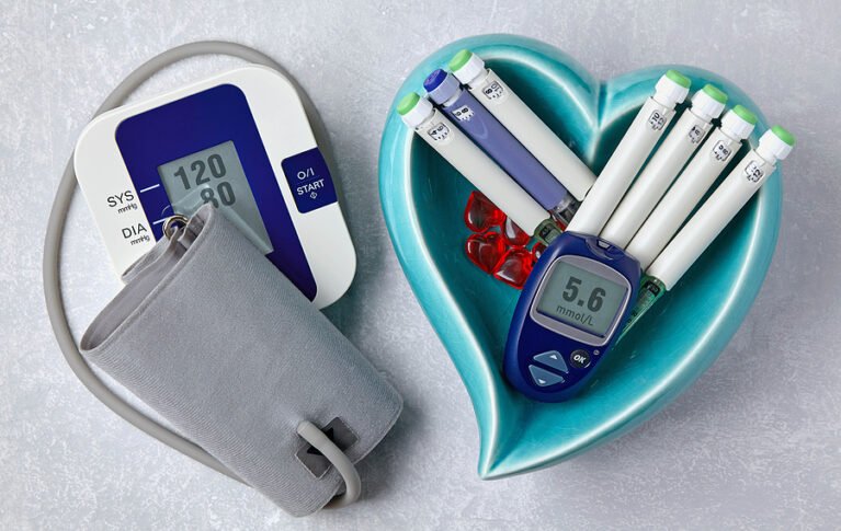 Blood pressure monitor, blood glucose monitor, insulin syringe pens in a blue heart-shaped bowl on a light gray concrete table. Medical equipment for health monitoring and therapy